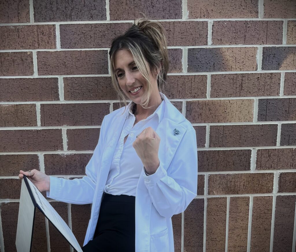 Dr. Brittney Ruedlinger stands in front of a brick wall, wearing a white lab coat and smiling while holding a certificate. She gestures with a fist pump of excitement and confidence.